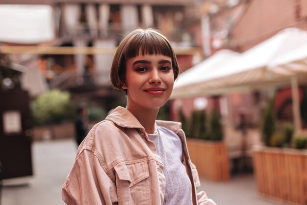 Charmante fille avec une coiffure courte en veste en jean beige sourire sincèrement en ville Femme positive aux cheveux bruns en vêtements légers posant dans la rue