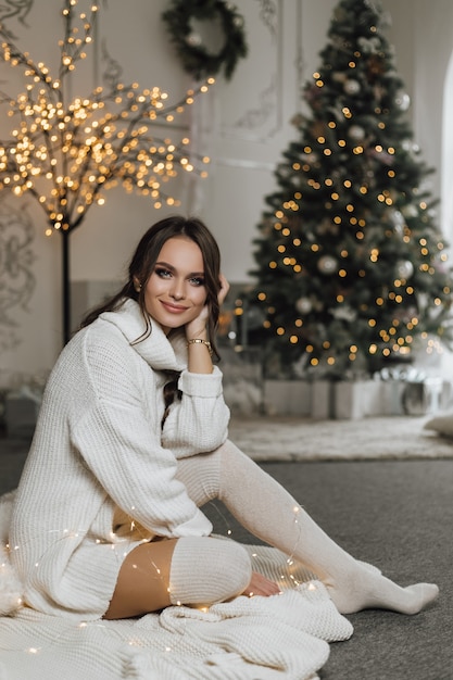 Charmante fille dans une robe tricotée a un look de rêve et est assise sur un fond d'arbre de Noël