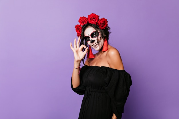 Charmante fille à l'image du squelette posant joyeusement. Portrait de jolie dame en haut noir avec des roses rouges en boucles montrant OK