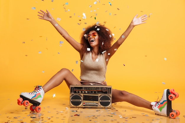 Photo gratuite charmante jeune femme africaine en vêtements rétro et scates à rouleaux jetant des confettis assis avec boombox