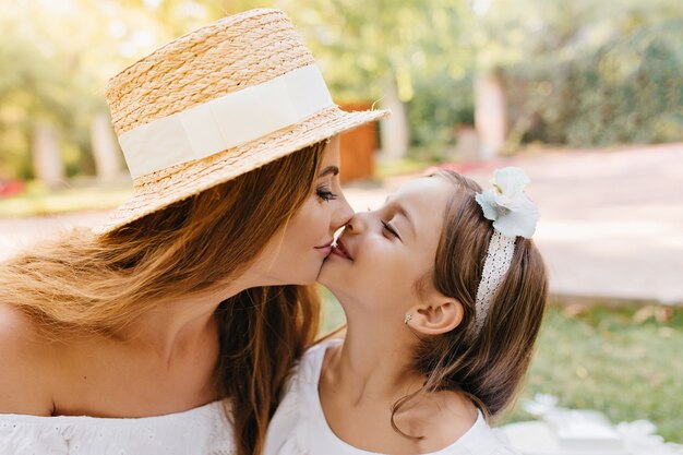 Charmante jeune femme aux longs cils noirs embrassant avec amour sa fille souriante. Portrait de gros plan de belle maman et mignonne femme aux cheveux longs avec ruban.