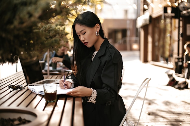 Charmante jeune femme brune en trench-coat noir prend des notes dans un cahier et travaille dans un ordinateur portable