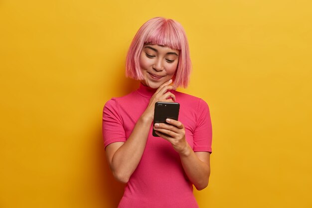 Charmante jeune femme avec une coiffure à la mode, sourit agréablement en regardant smartphone