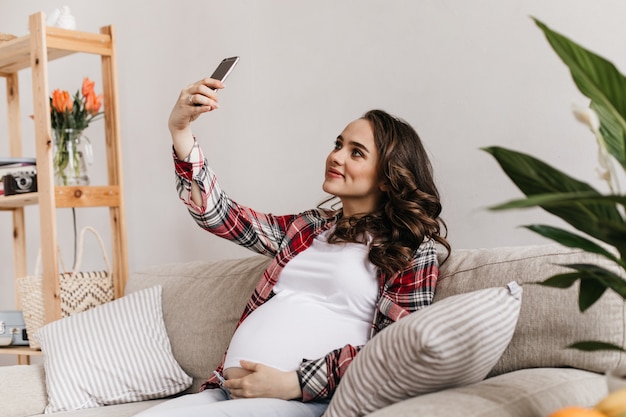 Charmante jeune femme enceinte en chemise à carreaux et tee-shirt blanc prend selfie et tient le téléphone