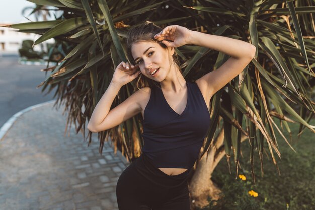 Charmante jeune femme étonnante en tenue de sport souriant sur arbre tropical. Étirement des mains, silhouette sexy, modèle sportif, humeur joyeuse, véritables émotions positives