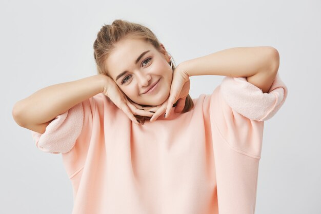 Charmante jeune femme européenne brillante vêtue de rose ayant des yeux bruns brillants et un large sourire à l'expression heureuse, tenant les mains sous son menton. Jolie étudiante posant à l'intérieur