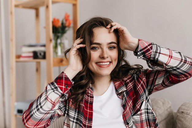 Charmante jeune femme frisée en chemise à carreaux et tee blanc sourit sincèrement Jolie fille brune se penche sur la caméra et pose dans le salon lumineux