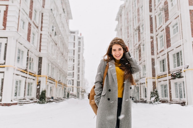 Charmante jeune femme en manteau aux longs cheveux bruns profitant des chutes de neige dans la grande ville. Émotions joyeuses, souriant, humeur de Noël, émotions de visage positif, temps d'hiver Place pour le texte.