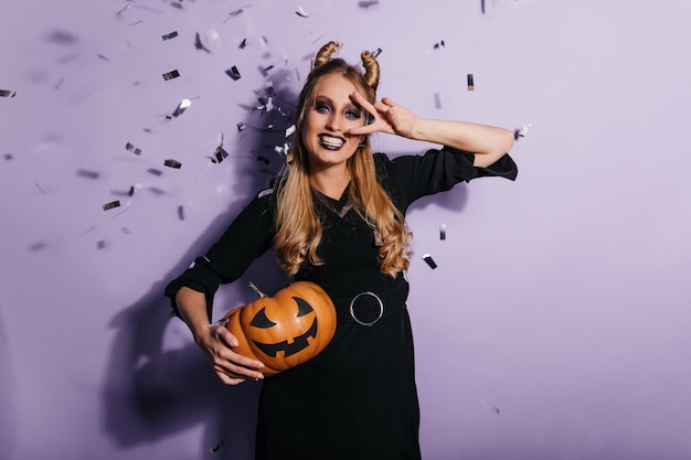 Charmante jeune femme en robe noire appréciant le carnaval d'halloween. photo de fille vampire souriante tenant une citrouille orange.