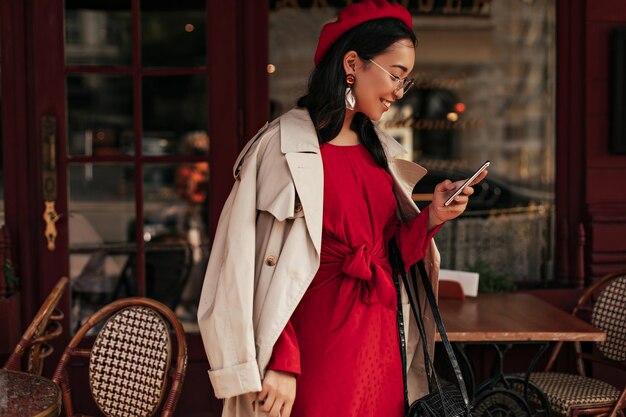 Charmante jeune femme en robe rouge béret et trench beige souriant et messagerie sur téléphone portable dans le café de la rue