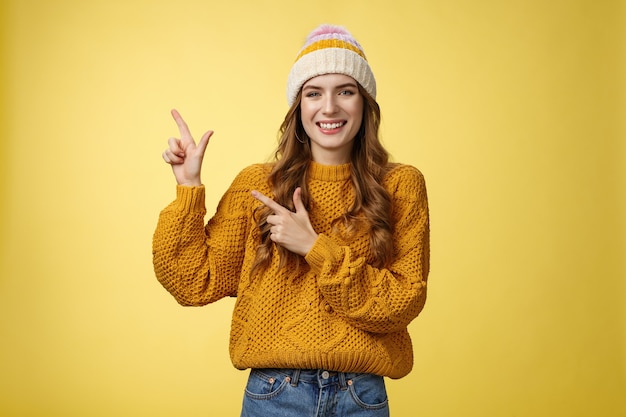 Charmante jeune femme souriante et insouciante portant un pull à chapeau pointant vers le coin supérieur gauche sur le côté partageant un lien intéressant une promotion géniale ravie de recommander un produit cool, fond jaune