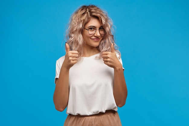 Photo gratuite charmante jeune hipster femme magnifique avec des cheveux rosés volumineux souriant joyeusement à la caméra, montrant le geste des pouces vers le haut