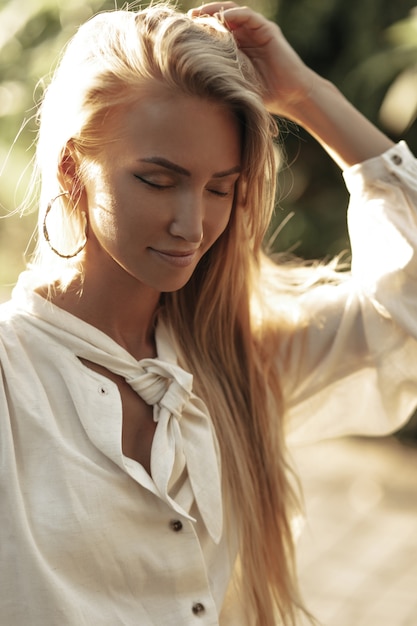 Photo gratuite charmante jolie femme bronzée en chemisier en coton blanc pose avec les yeux fermés à l'extérieur