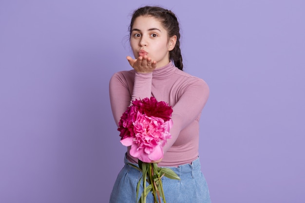 Charmante, jolie fille tenant le bouquet de pivoines dans les mains. Dame envoyant un baiser vexé posant isolé sur un mur lilas, fille avec des robes de tresses tenue décontractée.