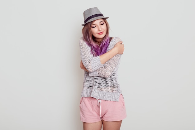 Photo gratuite charmante et magnifique femme européenne garde les yeux fermés, sourit de plaisir, se sent à l'aise, se serre dans ses bras, porte un chapeau, une chemise et un short, isolée sur fond gris.