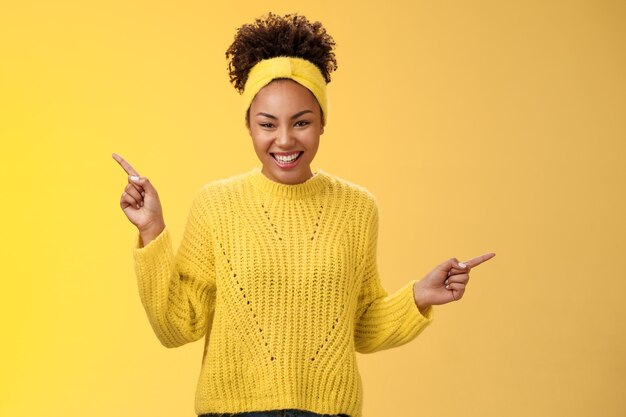 Charmante tendre romantique jeune femme afro-américaine des années 20 souriante amicale satisfaite riant belle caméra pointant vers le haut sur le côté montrer les choix de produits dans les deux sens, debout sur fond jaune.