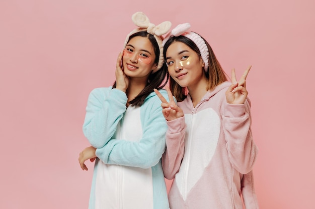 De charmantes femmes asiatiques en bandeaux de pyjama doux et avec des patchs pour les yeux sourient et montrent des vsigns sur fond rose