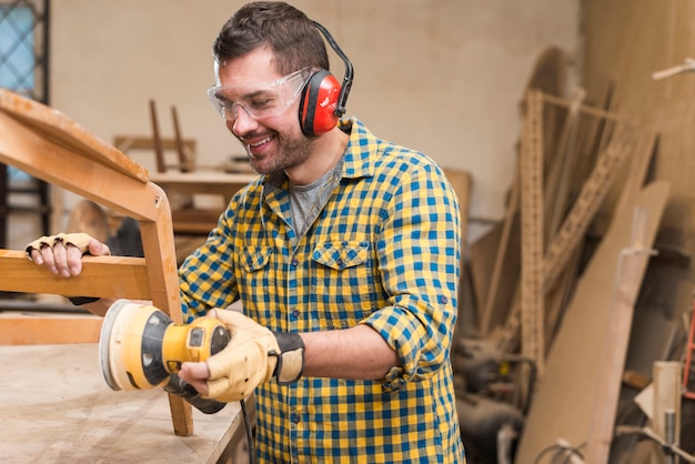 Charpentier souriant, ponçant un bois avec une ponceuse orbitale dans un établi