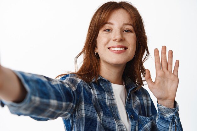 Chat vidéo de fille rousse moderne, agitant la main à l'appareil photo du smartphone et souriant, prenant selfie, enregistrement de blog sur téléphone debout sur blanc.