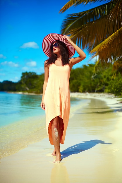 Chaude belle femme en chapeau de soleil coloré et robe marchant près de la plage océan sur une chaude journée d'été près de palm