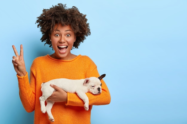 Cheerful Afro American woman fait un geste de paix, bénéficie d'une bonne journée, porte chiot endormi