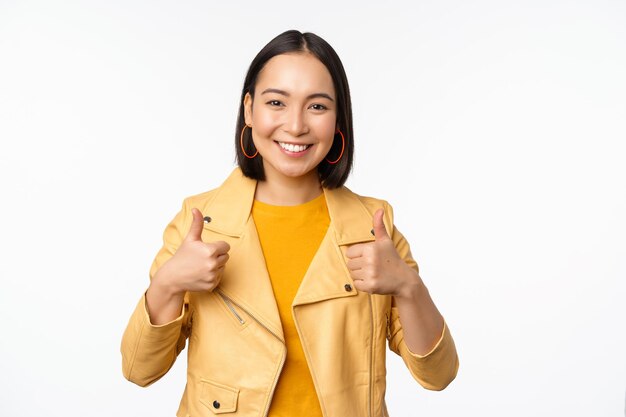 Cheerful asian woman smiling heureux montrant les pouces vers le haut dans l'approbation debout dans des vêtements décontractés sur fond blanc