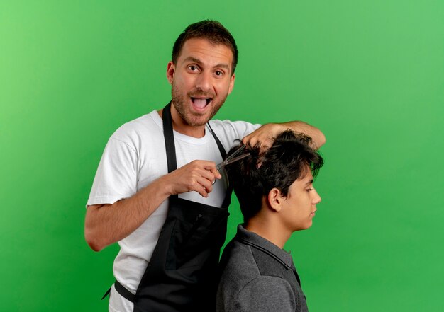 Cheerful Barber Man In Tablier Couper Les Cheveux Avec Des Ciseaux De Client Satisfait Debout Sur Mur Vert