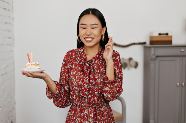 Cheerful brunette Asian woman in floral dress croise les doigts et sourit largement Happy young girl détient morceau de gâteau d'anniversaire et fait voeu