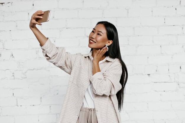 Cheerful brunette woman in beige cardigan sourit sincèrement sur fond de mur de briques blanches Dame bronzée de bonne humeur prend selfie
