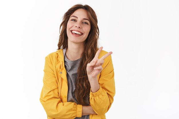 Cheerful enthousiaste femme magnifique coiffure châtaigne frisée spectacle geste de victoire souriant rire insouciant ayant une humeur optimiste optimiste debout mur blanc profiter de l'été