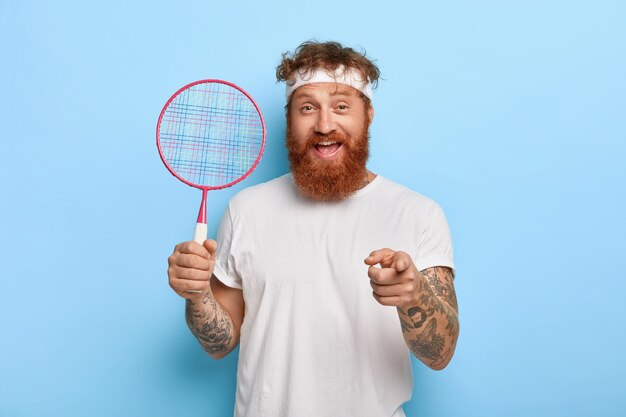 Cheerful friendly joueur de tennis aux cheveux rouges tient la raquette tout en posant contre le mur bleu