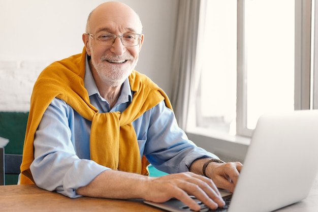 Cheerful Mature Homme Chauve Barbu Portant Des Lunettes Et Un Pull Sur Chemise Formelle Bleue Souriant Joyeusement Tout En Tapant Sur Un Ordinateur Portable, Jouer à Des Jeux Vidéo Pendant La Pause Déjeuner