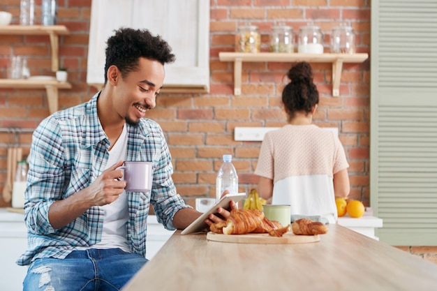 Cheerful Mixed Race Male Regarde La Comédie Sur Tablette, Utilise Une Connexion Internet Gratuite, Boit Du Café