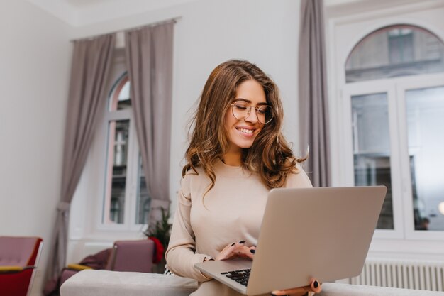 Cheerful smart lady dans des verres travaillant avec un ordinateur portable dans une chambre confortable