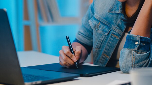 Cheerful young graphic designer lady using digital graphic tablet while working late in modern office at night, Asia professional female using laptop computer retoucher sitting in living room at home.