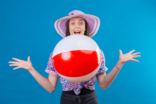 Cheerful young woman in summer hat portant lancer ballon gonflable souriant avec visage heureux debout sur l'espace bleu