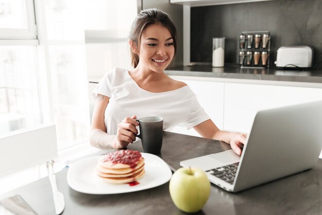 Cheerful young woman sitting at the kitchen using laptop