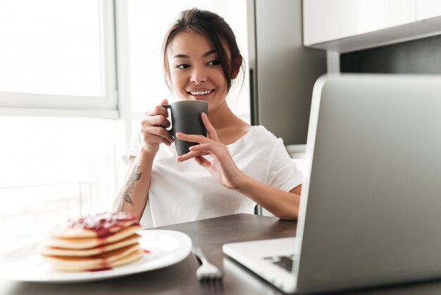 Cheerful young woman sitting at the kitchen using laptop