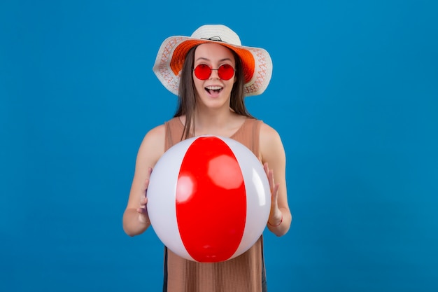 Cheerful young woman with hat portant des lunettes de soleil rouges tenant ballon gonflable avec sourire sur le visage debout sur bleu