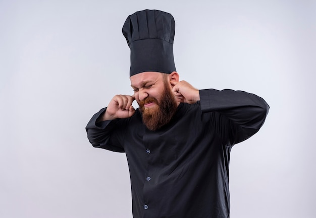 Photo gratuite un chef barbu en colère homme en uniforme noir tenant les oreilles avec les doigts sur un mur blanc