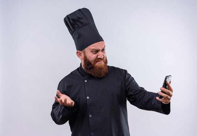 Un chef barbu confus en uniforme noir montrant une expression de colère tout en regardant son téléphone portable sur un mur blanc