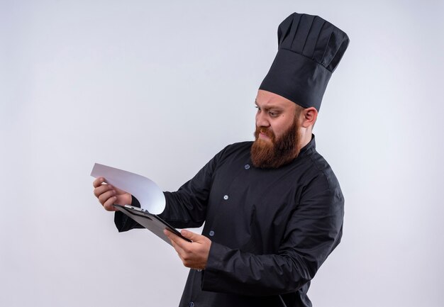 Un chef barbu confus en uniforme noir regardant un dossier vierge avec une expression agressive sur un mur blanc