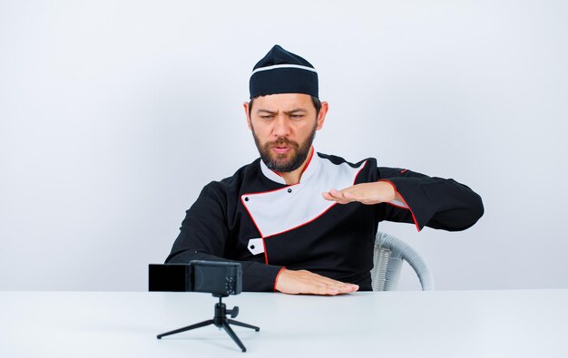 Le chef blogueur montre un geste de taille en s'asseyant devant sa mini caméra sur fond blanc