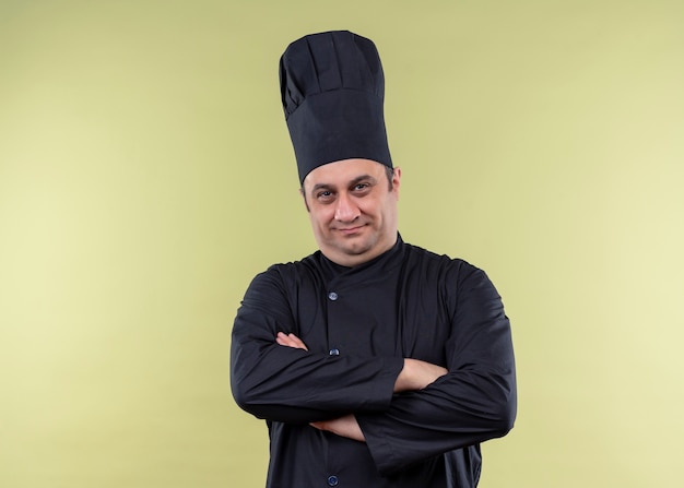 Chef cuisinier mâle portant l'uniforme noir et chapeau de cuisinier à la caméra avec les mains croisées à la confiance debout sur fond vert