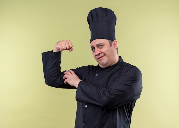 Chef cuisinier mâle portant l'uniforme noir et chapeau de cuisinier levant le poing montrant les biceps à la confiance debout sur fond vert