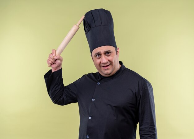 Chef cuisinier mâle portant l'uniforme noir et chapeau de cuisinier se gratter la tête avec la broche de roulement à la confusion debout sur fond vert