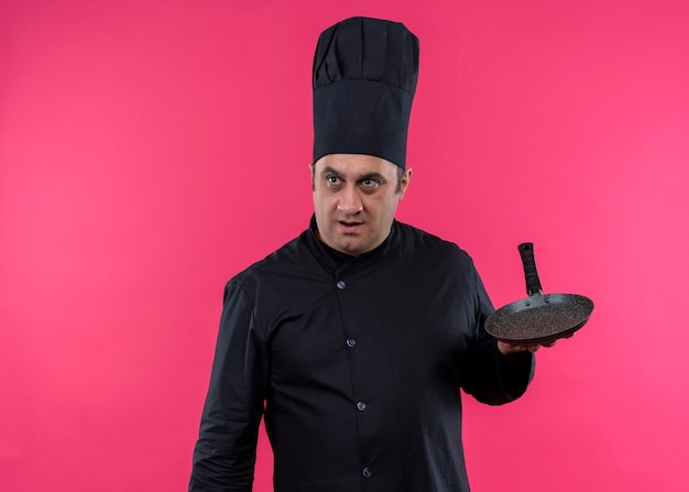 Chef cuisinier mâle portant l'uniforme noir et chapeau de cuisinier tenant une casserole à côté surpris debout sur fond rose