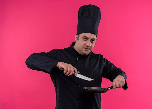 Chef cuisinier mâle portant l'uniforme noir et chapeau de cuisinier tenant la casserole et le couteau regardant la caméra avec un visage sérieux debout sur fond rose