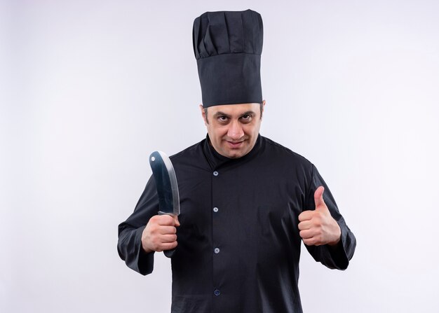 Chef cuisinier mâle portant l'uniforme noir et chapeau de cuisinier tenant un couteau de cuisine pointu souriant montrant les pouces vers le haut debout sur fond blanc