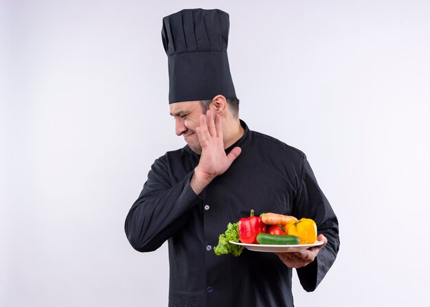 Chef cuisinier mâle portant l'uniforme noir et cook hat holding plaque avec des légumes frais à côté avec une expression dégoûtée debout sur fond blanc